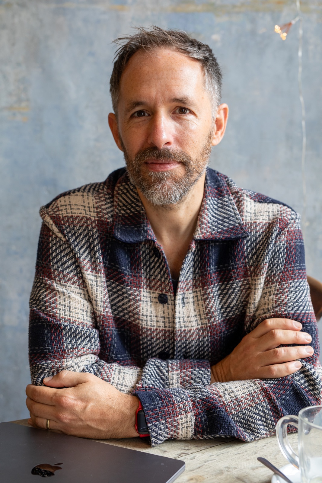 Simon Booth-Lucking sitting at a café table in a woven check jacket, a closed laptop in front of him, against a peeling blue wall.
